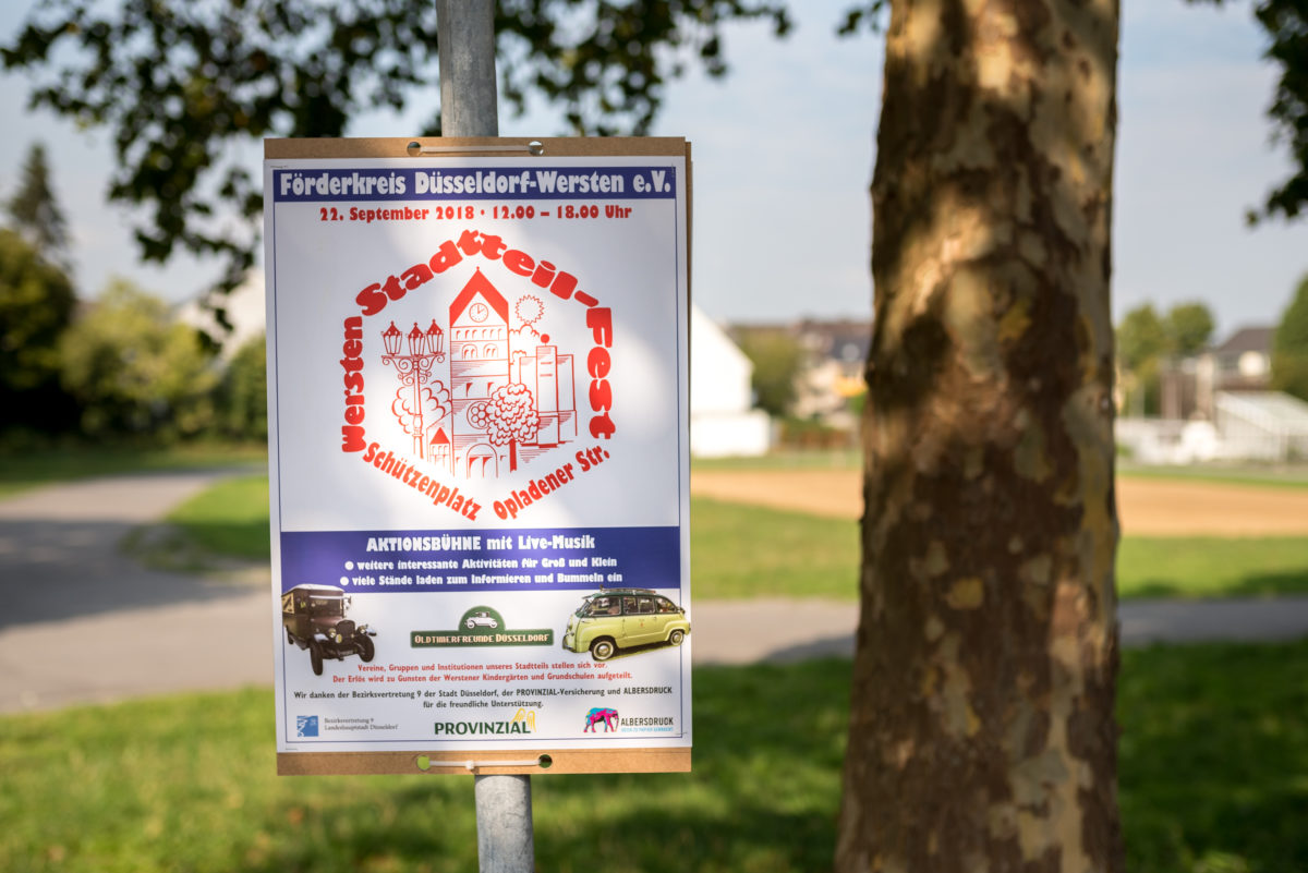 Ein Plakat hängt an einem Pfahl. Dort ist die Einladung zum Stadtteilfest zu erkennen. Im Hintergrund ist der (noch) leere Schützenplatz zu sehen.